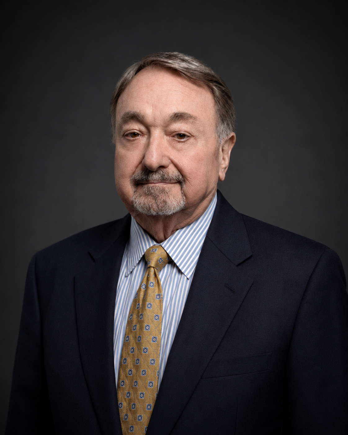 Head-and-shoulders portrait of an older man with gray hair and a goatee, wearing a navy blazer, blue-striped shirt, and yellow patterned tie against a dark studio backdrop.